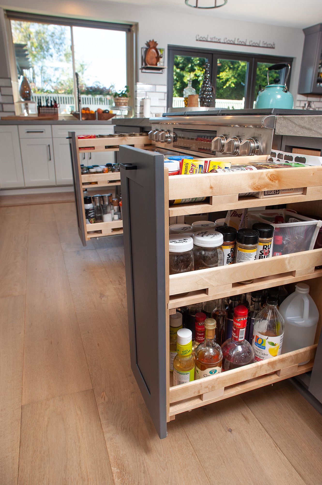 Gray kitchen cabinet with pull-out shelves, filled with spices, jars, and bottles, set on wood flooring.