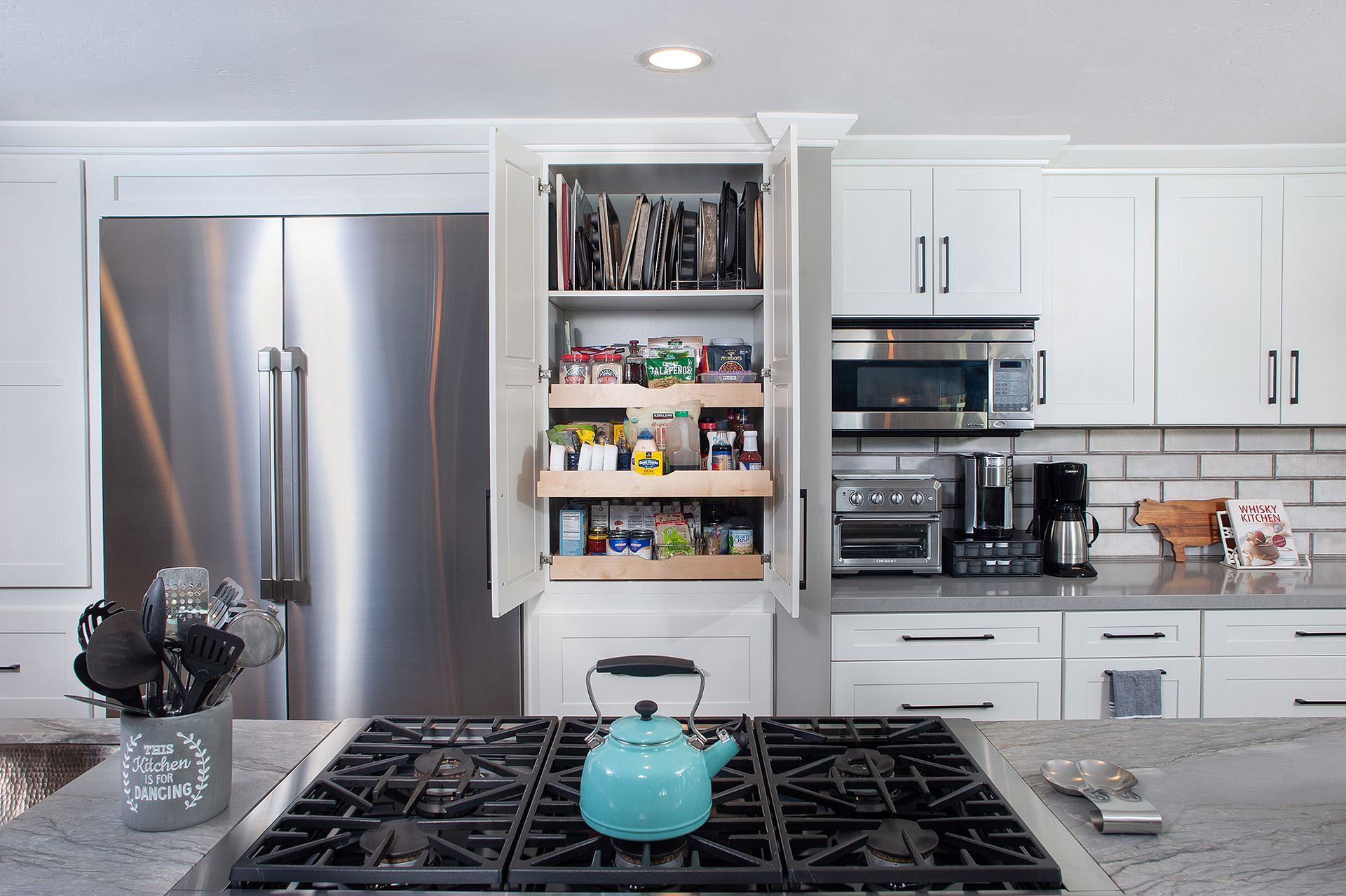 A bright kitchen with a stainless steel refrigerator, open pantry with pull-out shelves, and gas stove.