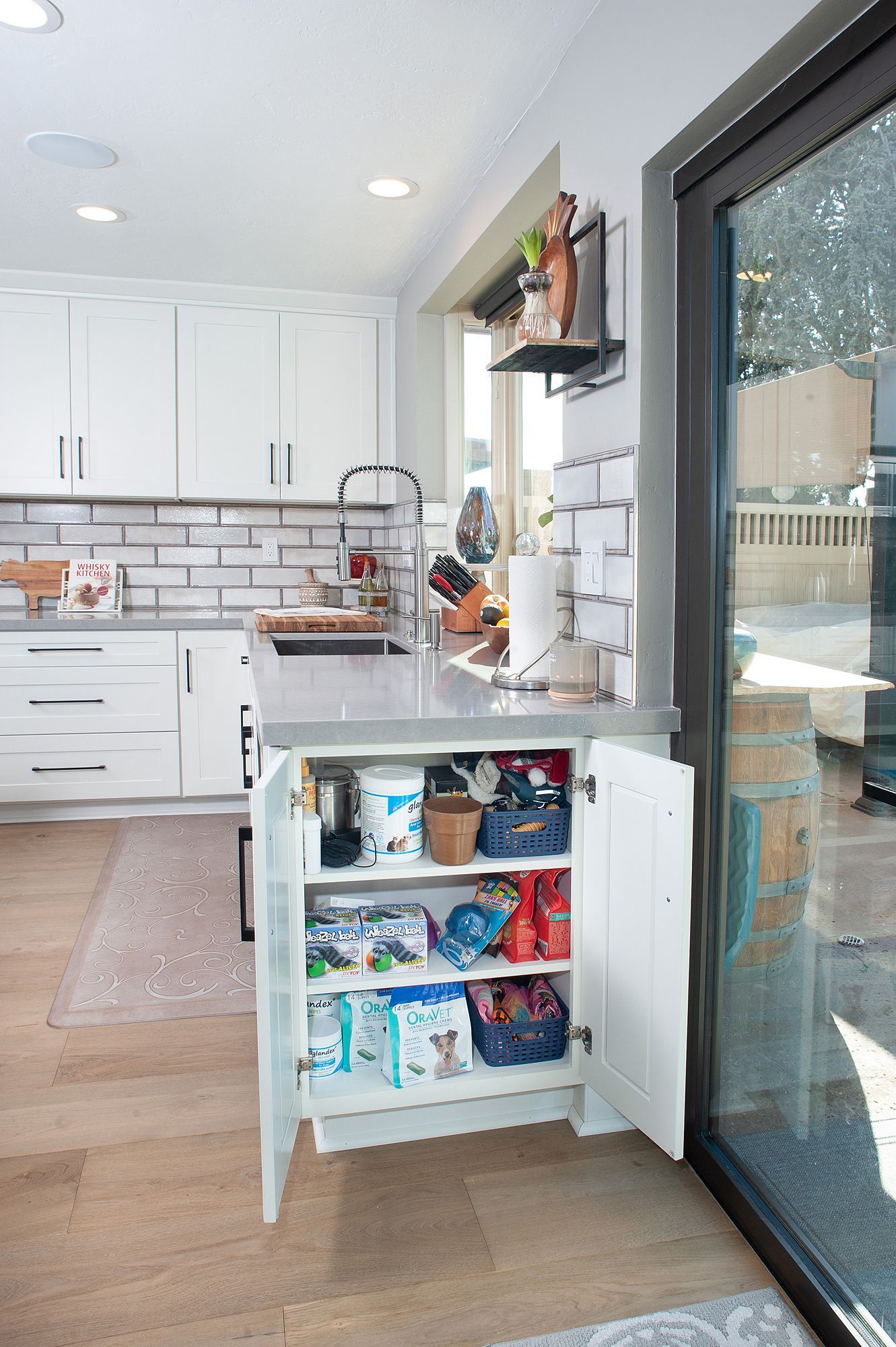 White kitchen with open cabinet revealing snacks and storage. Near window, light wood floors.
