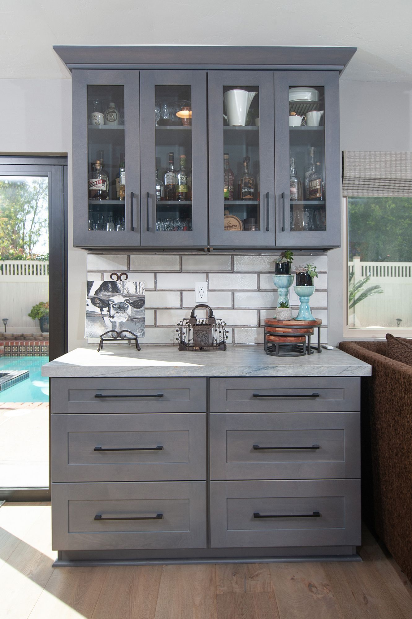 Gray cabinet bar with glass upper doors, stacked on drawers, set against a brick backsplash, near a pool.