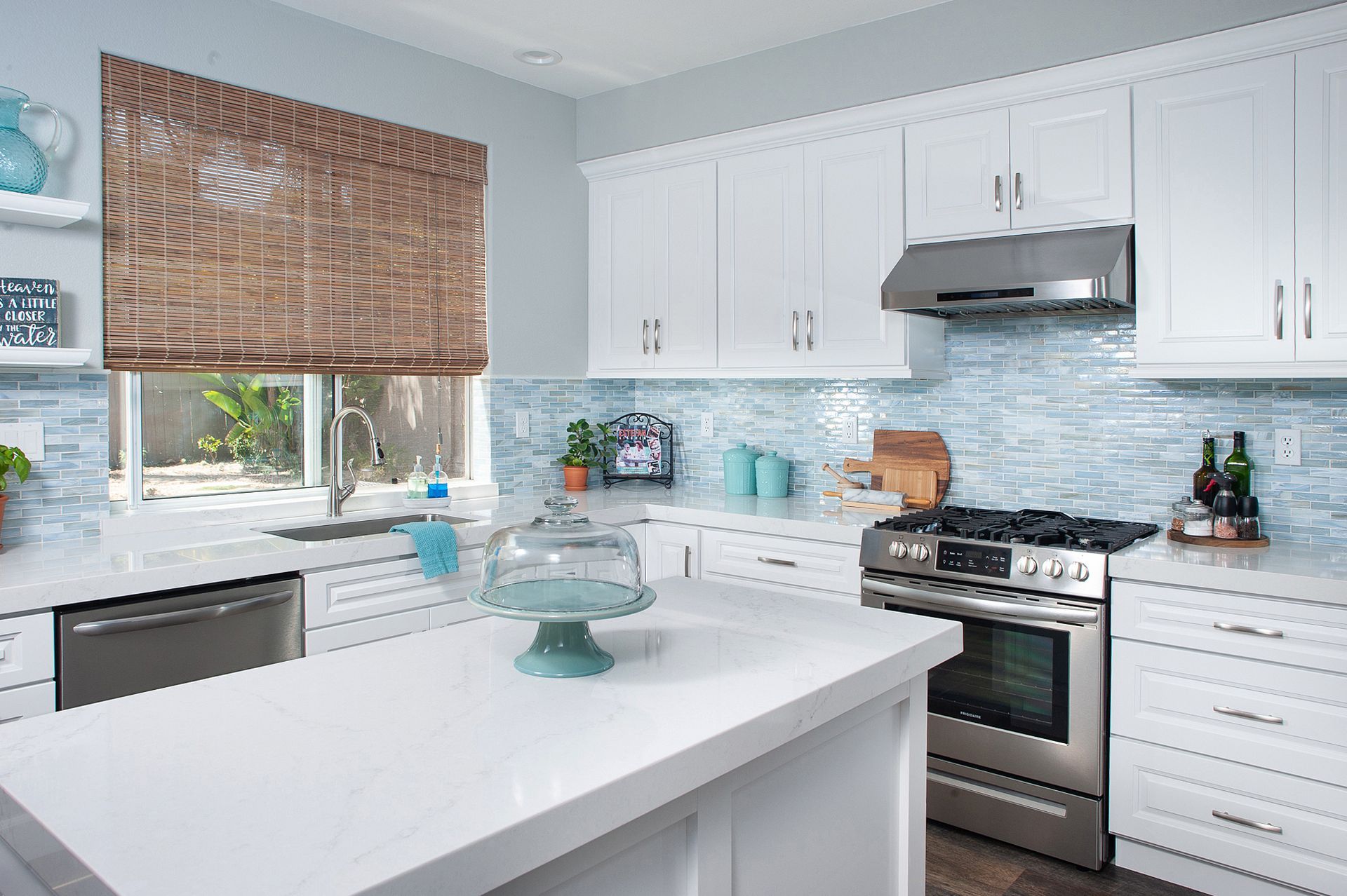 White kitchen with island, stainless steel appliances, blue tile backsplash, window with bamboo shade.