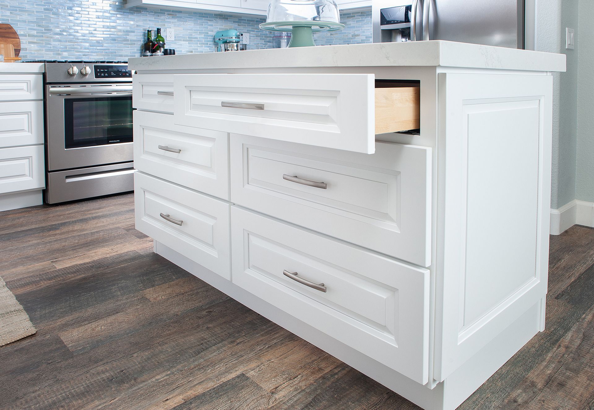 White kitchen island with drawers, one open, near a stainless steel oven on a wood floor.