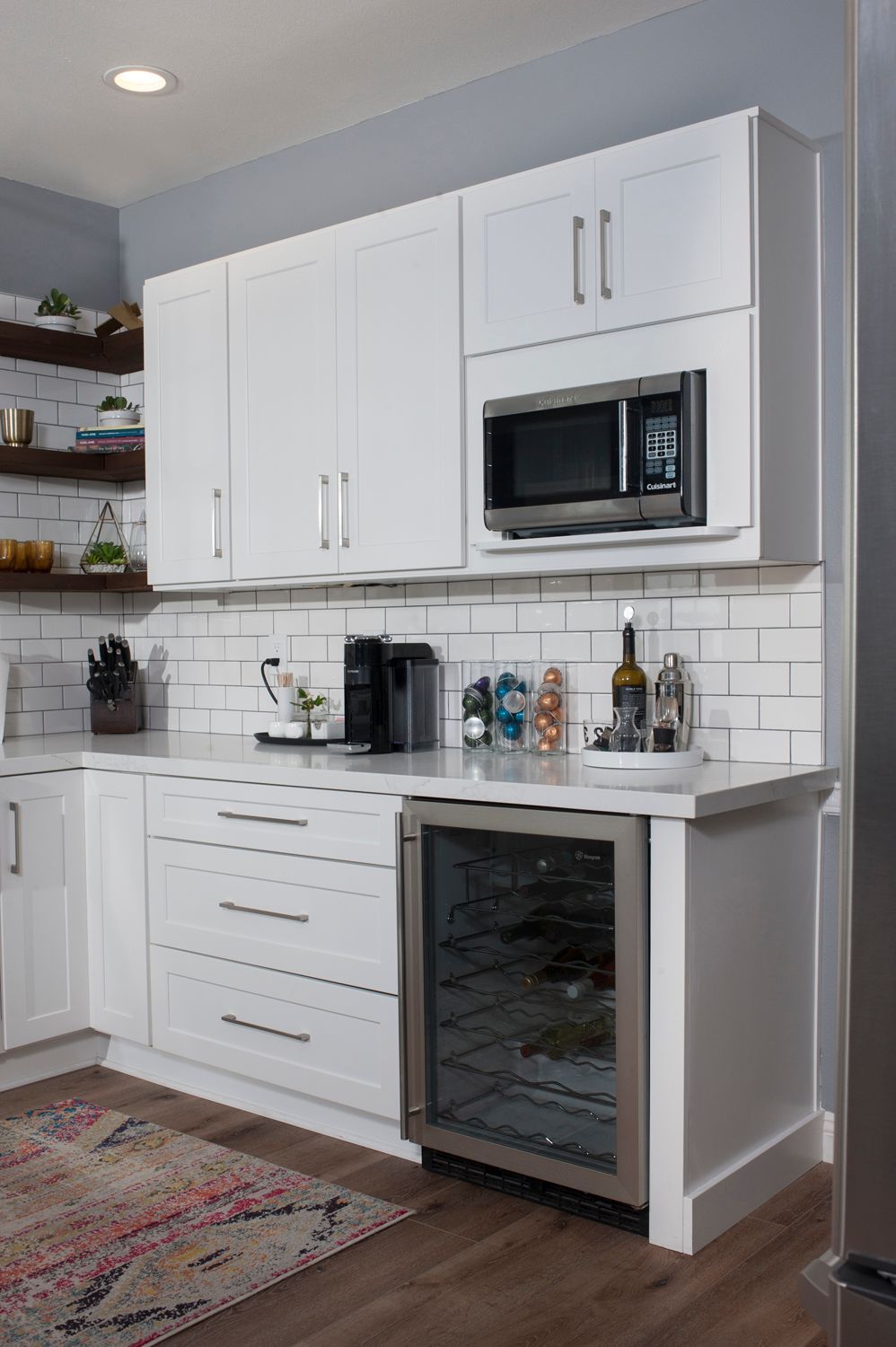White kitchen with wine fridge, microwave, and cabinetry. Subway tile backsplash, wooden floor, and rug.
