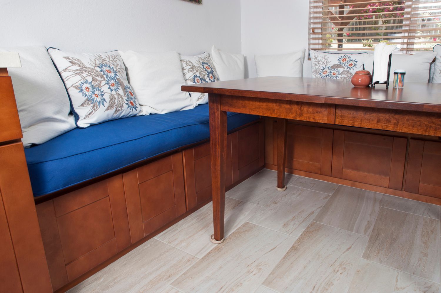 A built-in dining nook with a wooden table and storage bench, blue cushions, white pillows, and a window.