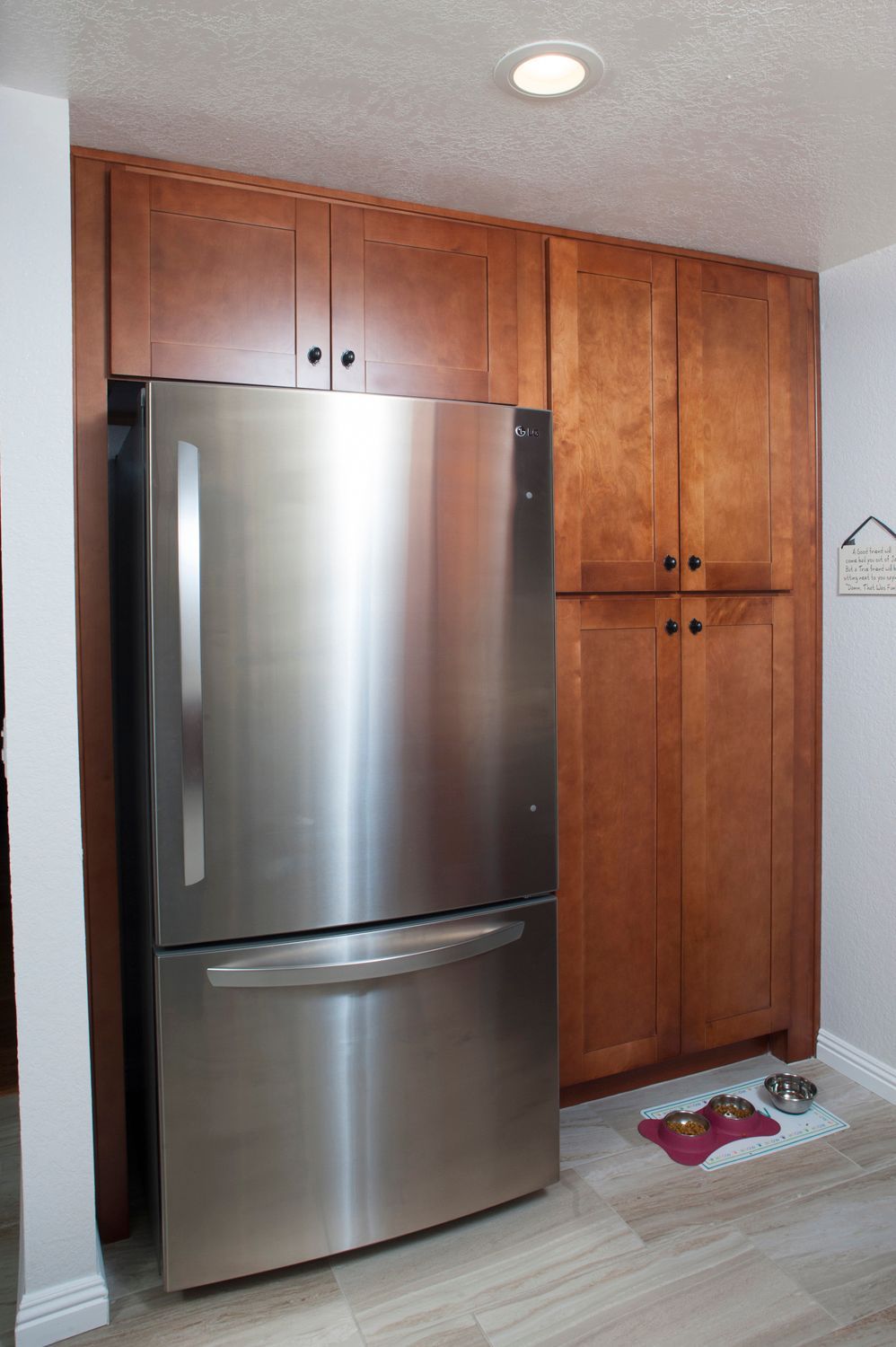 Stainless steel refrigerator next to tall wooden cabinets. Tile floor, dog bowl.