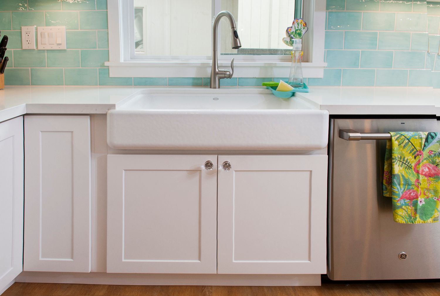 White kitchen sink with cabinet doors and a stainless steel dishwasher. Turquoise tile backsplash.