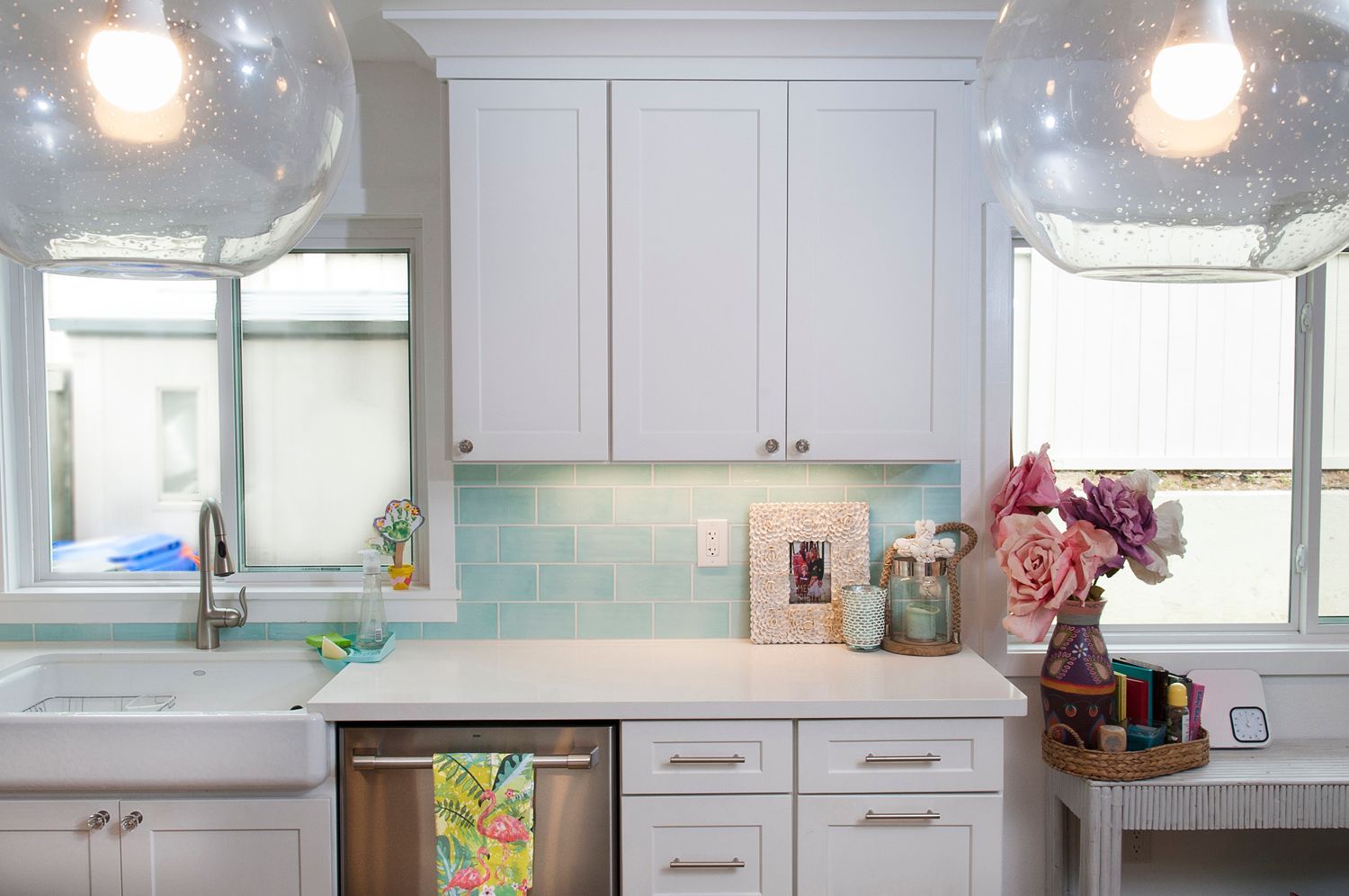 White kitchen cabinets with blue backsplash, sink, and windows; decorated with flowers and a photo.