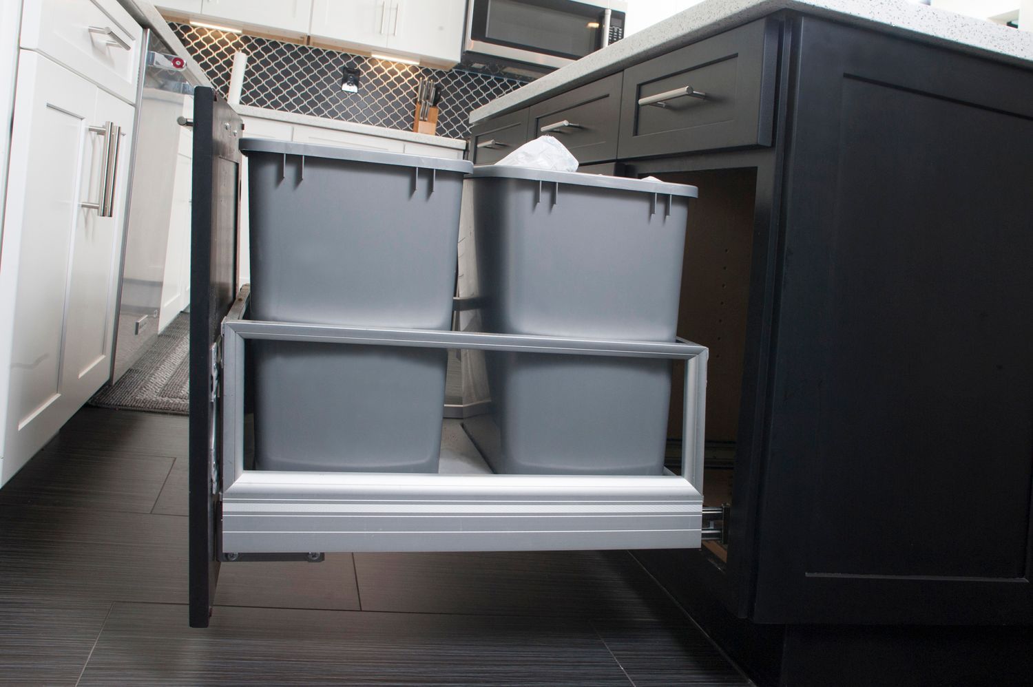 Pull-out kitchen cabinet with four gray trash bins, in a black cabinet.