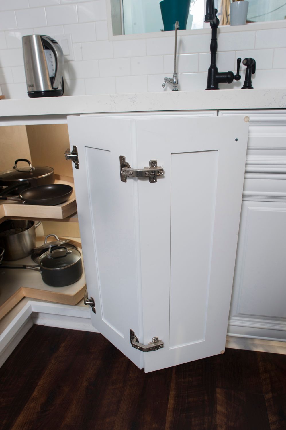 White kitchen cabinet door open, revealing pots and pans, dark wood floor.