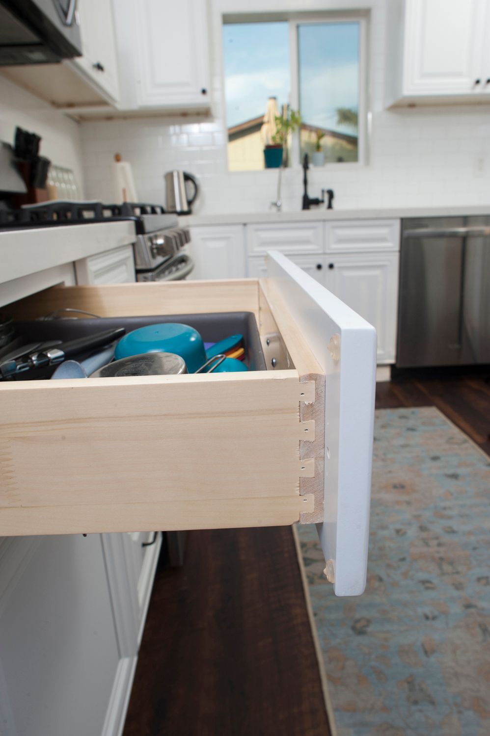 Open kitchen drawer with dovetail joints, filled with utensils. White cabinets, stainless steel appliances, and blue rug.