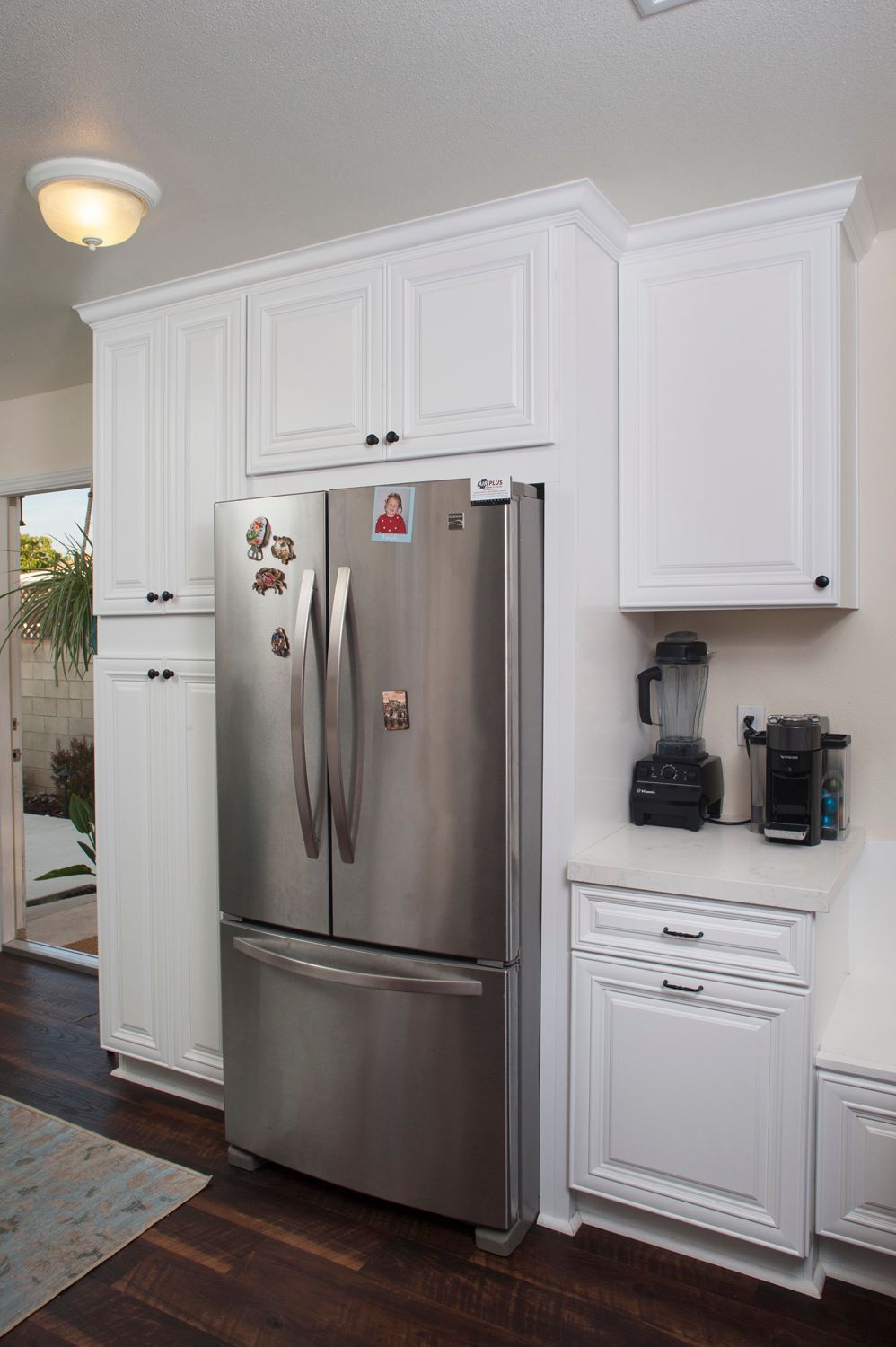 White kitchen cabinets surround a stainless steel refrigerator. Blender and coffee maker sit on a countertop.