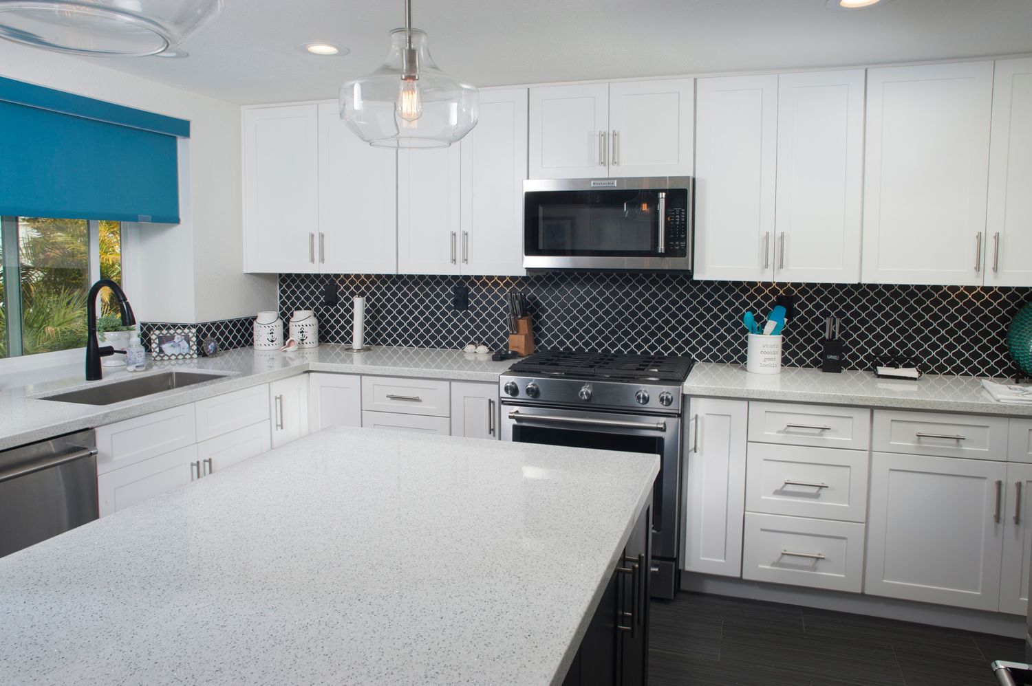 Modern white kitchen with quartz countertops, black backsplash, stainless steel appliances, and turquoise window shade.