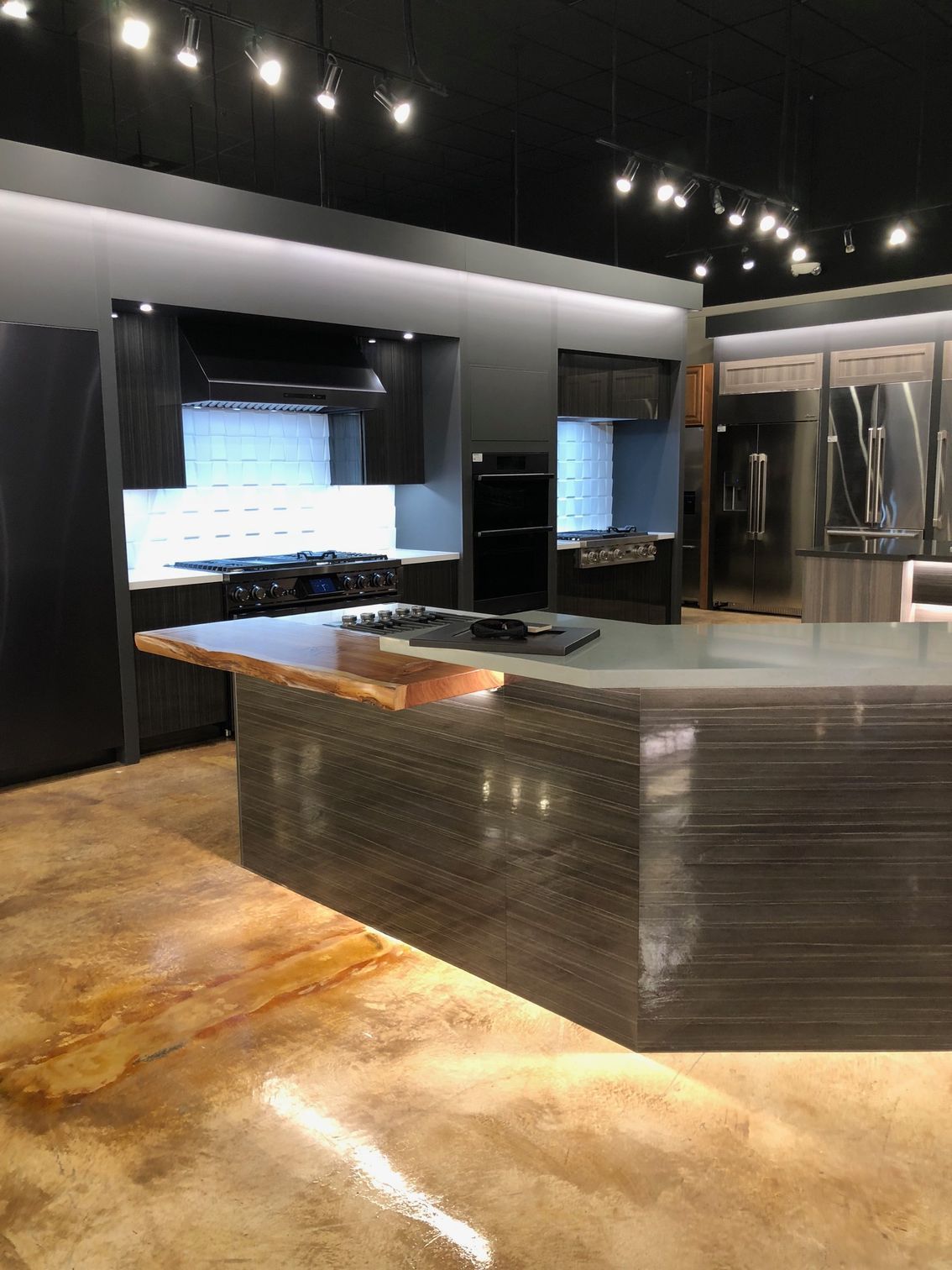 Modern kitchen display with a large island and dark cabinetry. Polished concrete floor.