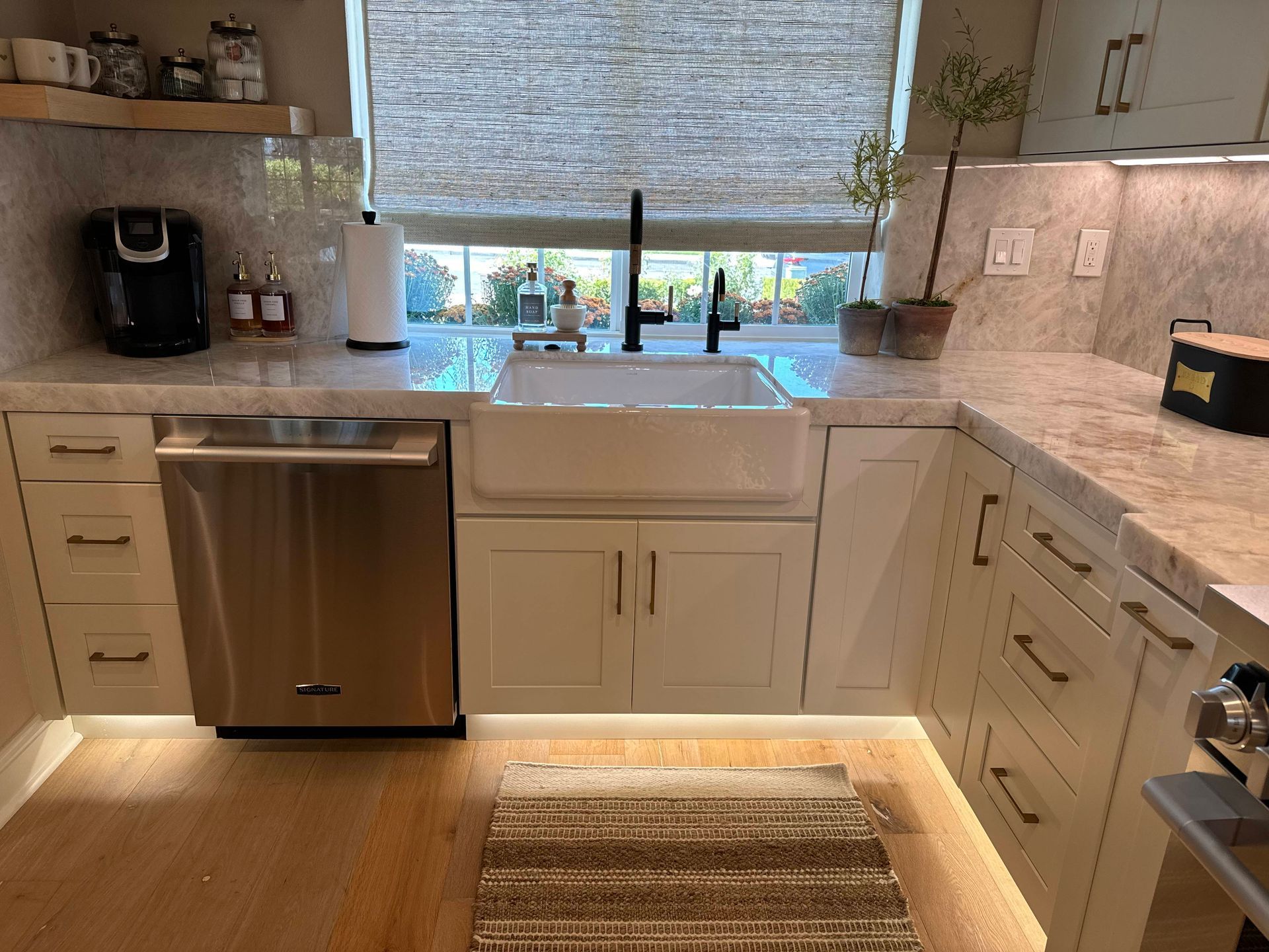 Kitchen with farmhouse sink, stainless steel dishwasher, beige cabinets, and a window with a woven shade.