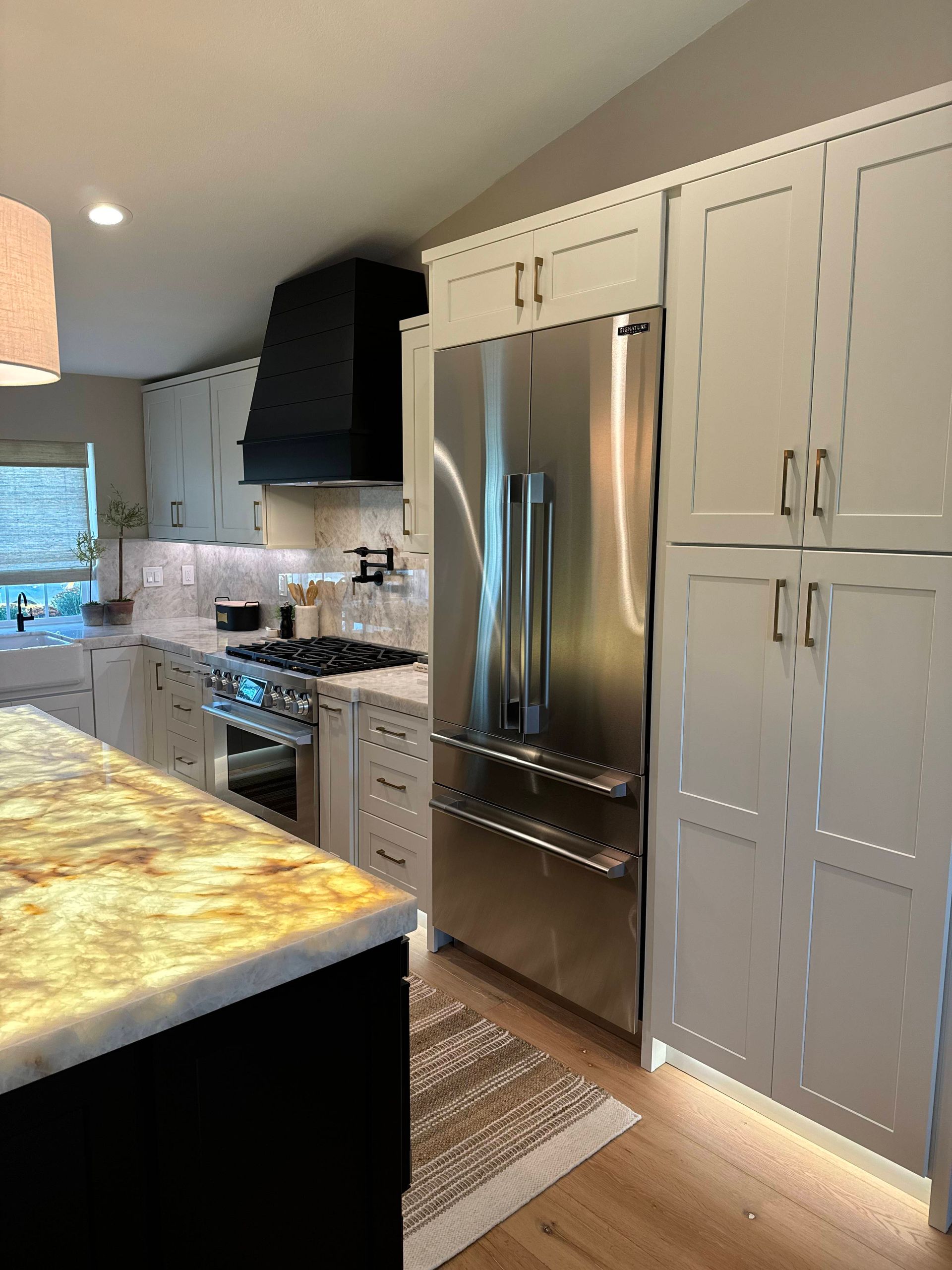 Modern kitchen with stainless steel refrigerator, white cabinets, black range hood, and marble countertops.