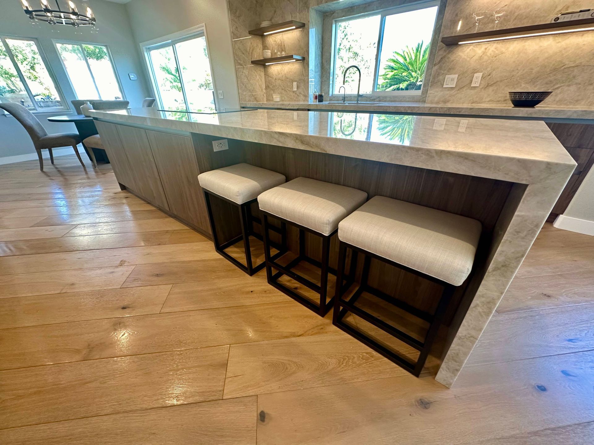 Kitchen island with three white-cushioned bar stools, light wood floors, and stone countertops.
