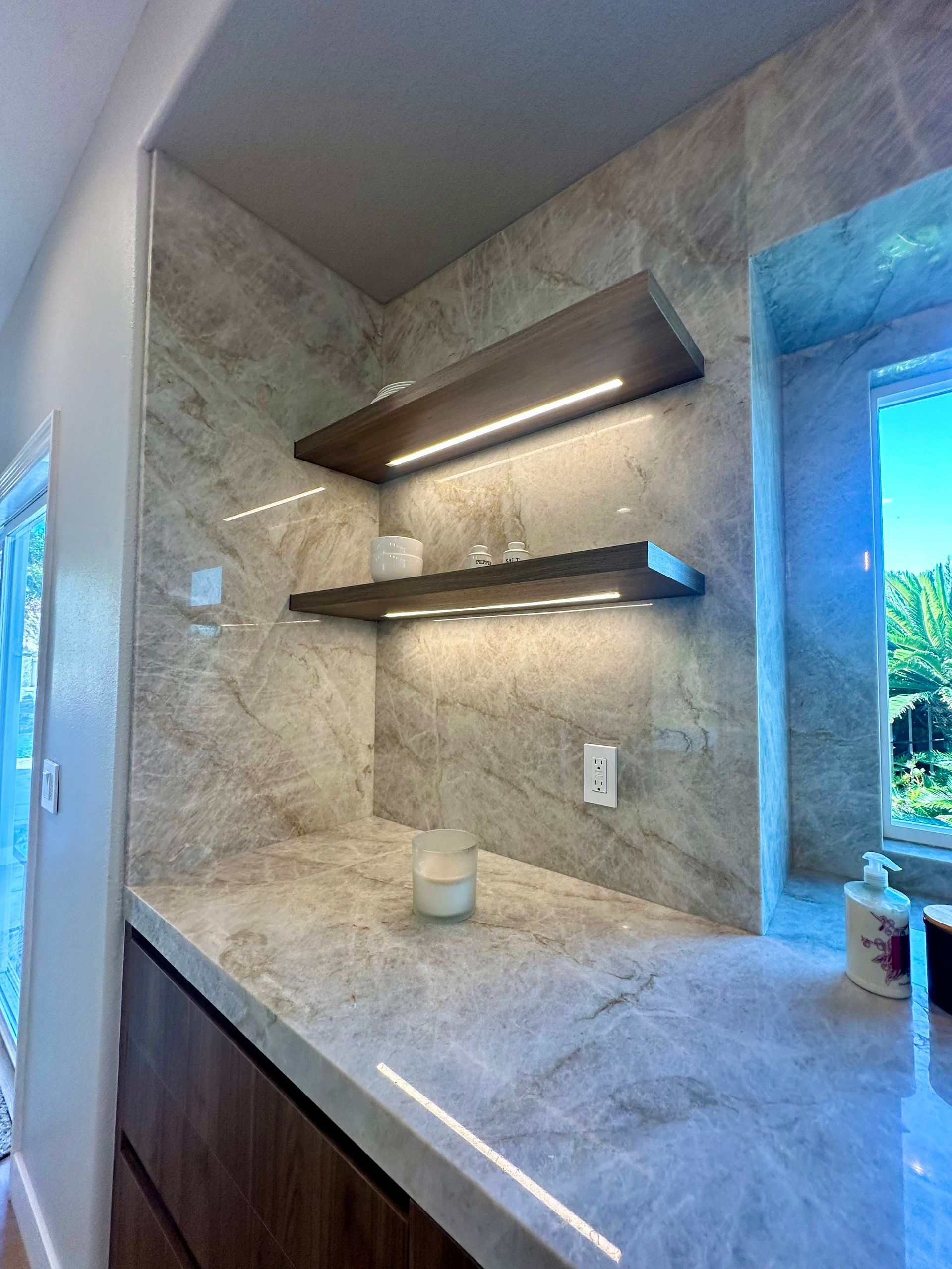 Built-in shelves with LED lighting in a kitchen nook. Gray marble-like backsplash, dark wood shelves, and a window.