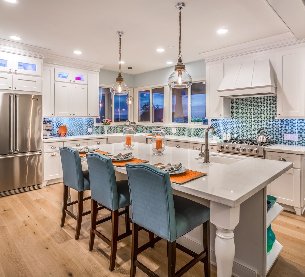 White kitchen with an island, blue chairs, wood floor, and blue/white backsplash.