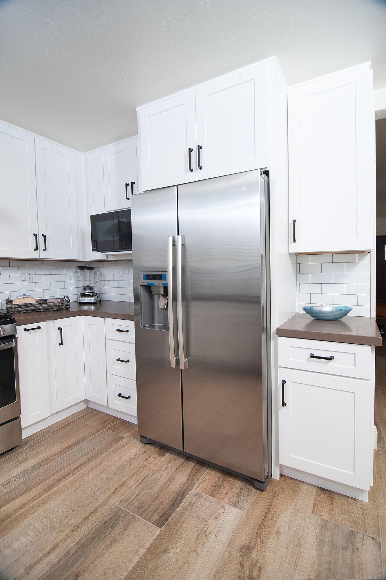 Stainless steel refrigerator in white kitchen with cabinets, wood floors, and brown countertops.