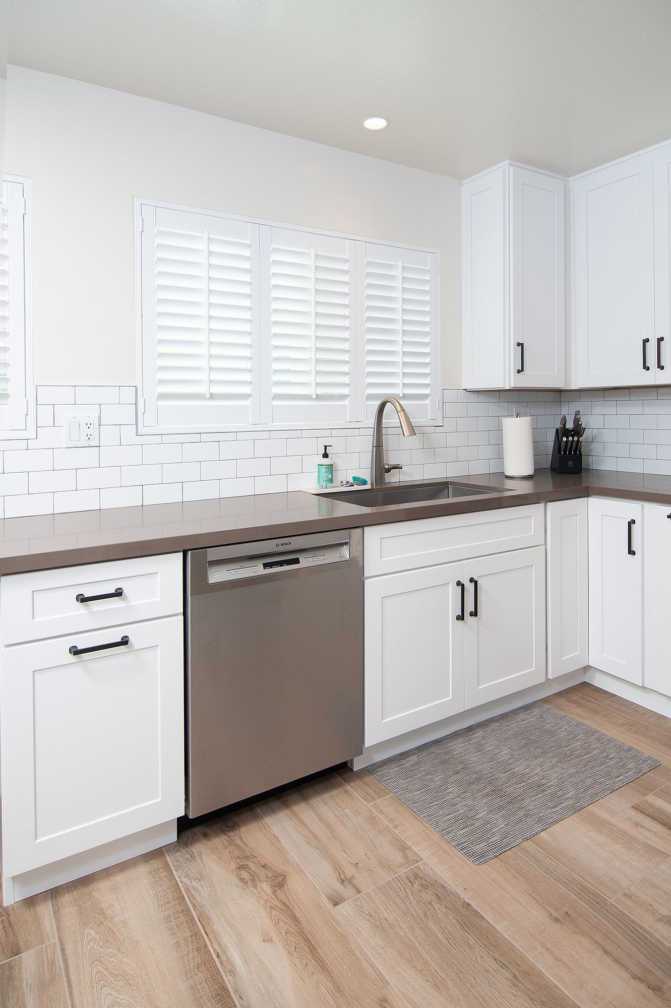 White kitchen with stainless steel dishwasher, cabinets, and gray countertops. Wood floors.
