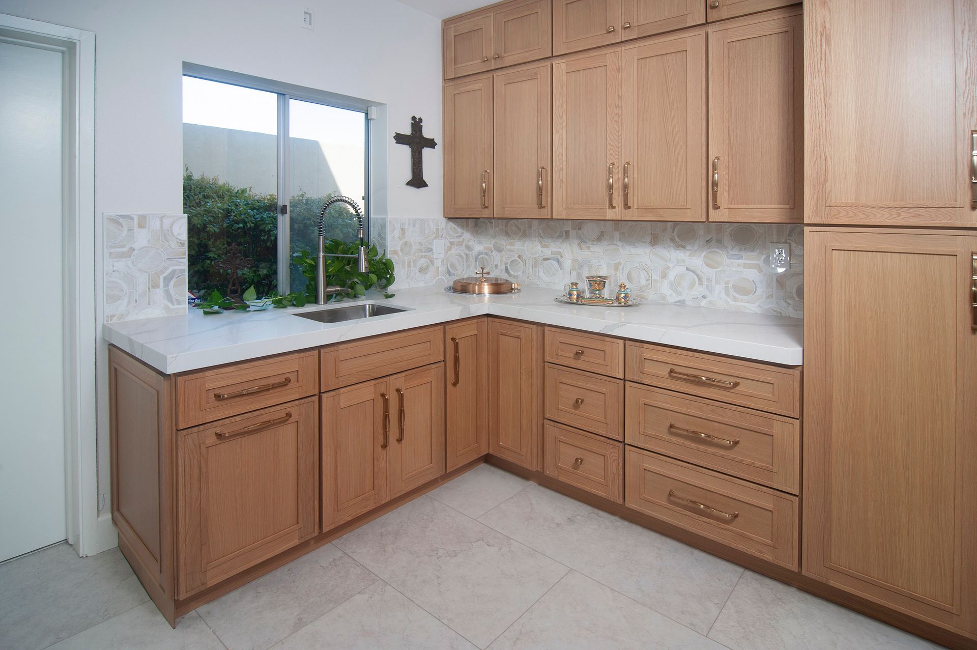 Light-wood kitchen cabinets, white countertop, window over sink, cross on wall.