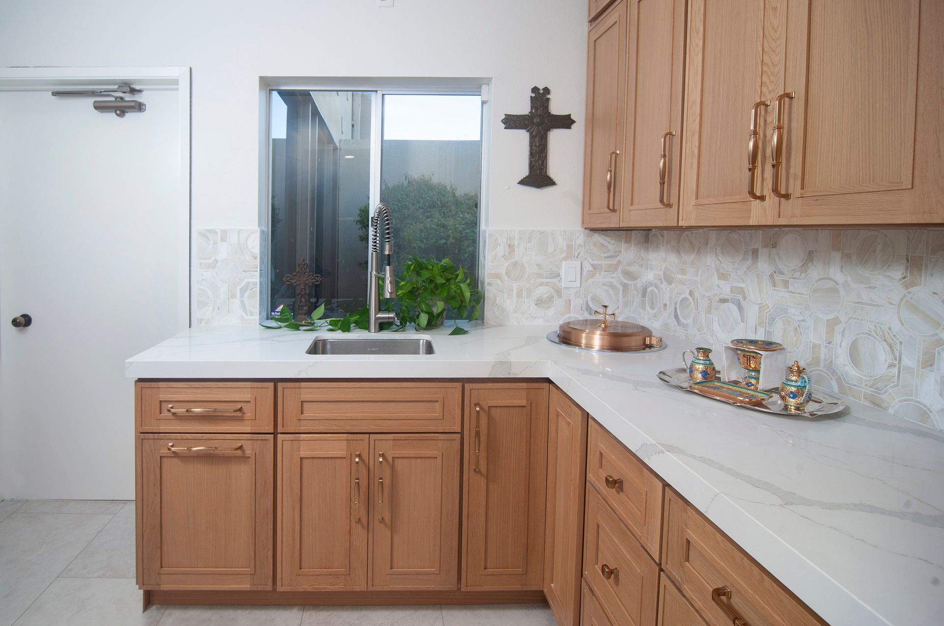 Light oak cabinets and white countertop in a kitchen with a window and cross decoration.