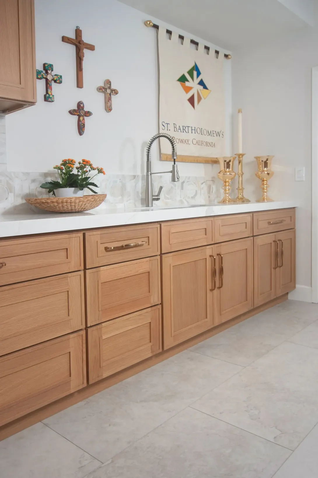 Light wood kitchen cabinets with white countertop, sink, crucifixes, and gold faucet against a white wall