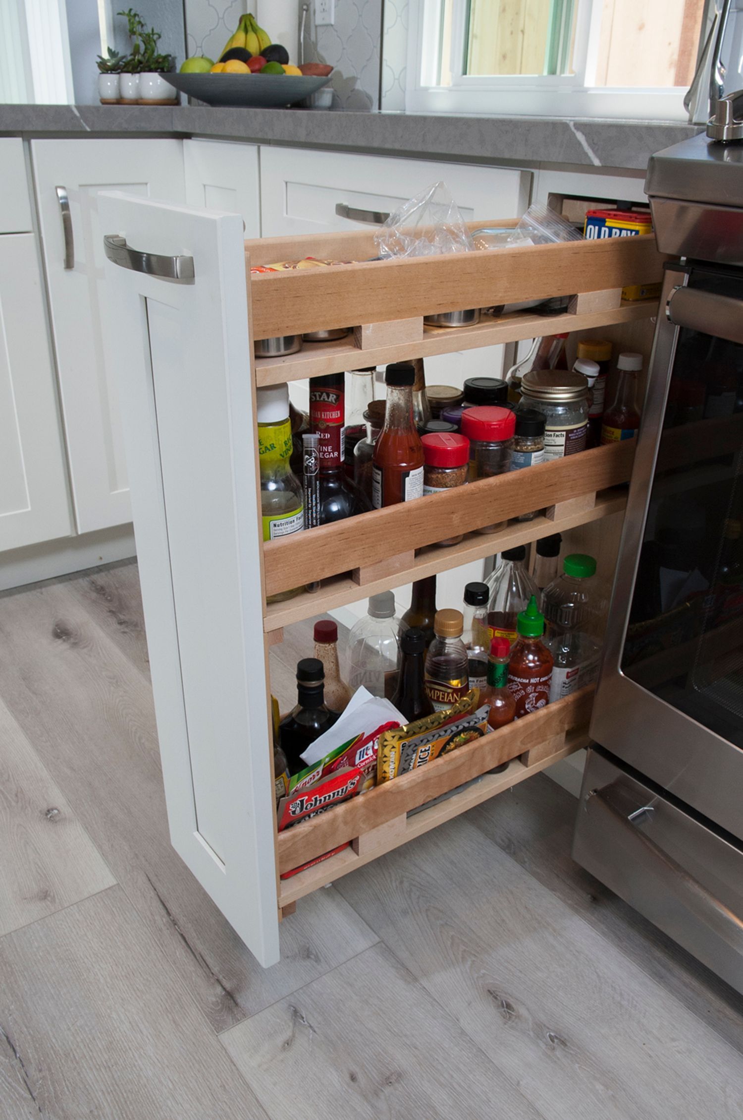 Pull-out kitchen cabinet with spices, sauces, and snacks. Light wood shelves, white cabinet door, near a stove.