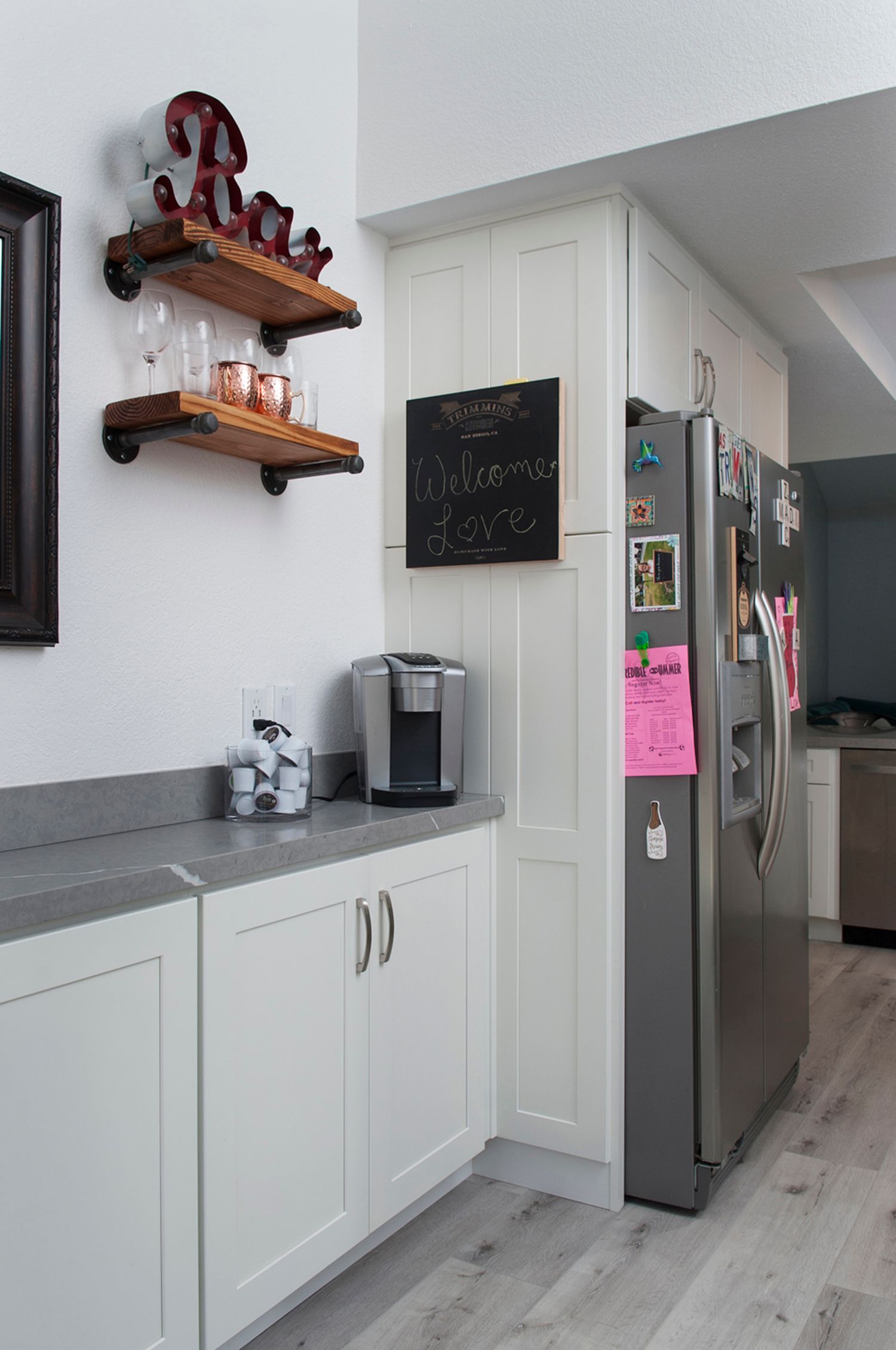 A kitchen with white cabinets, a gray countertop, and a stainless steel refrigerator. Wooden shelves hold glassware.