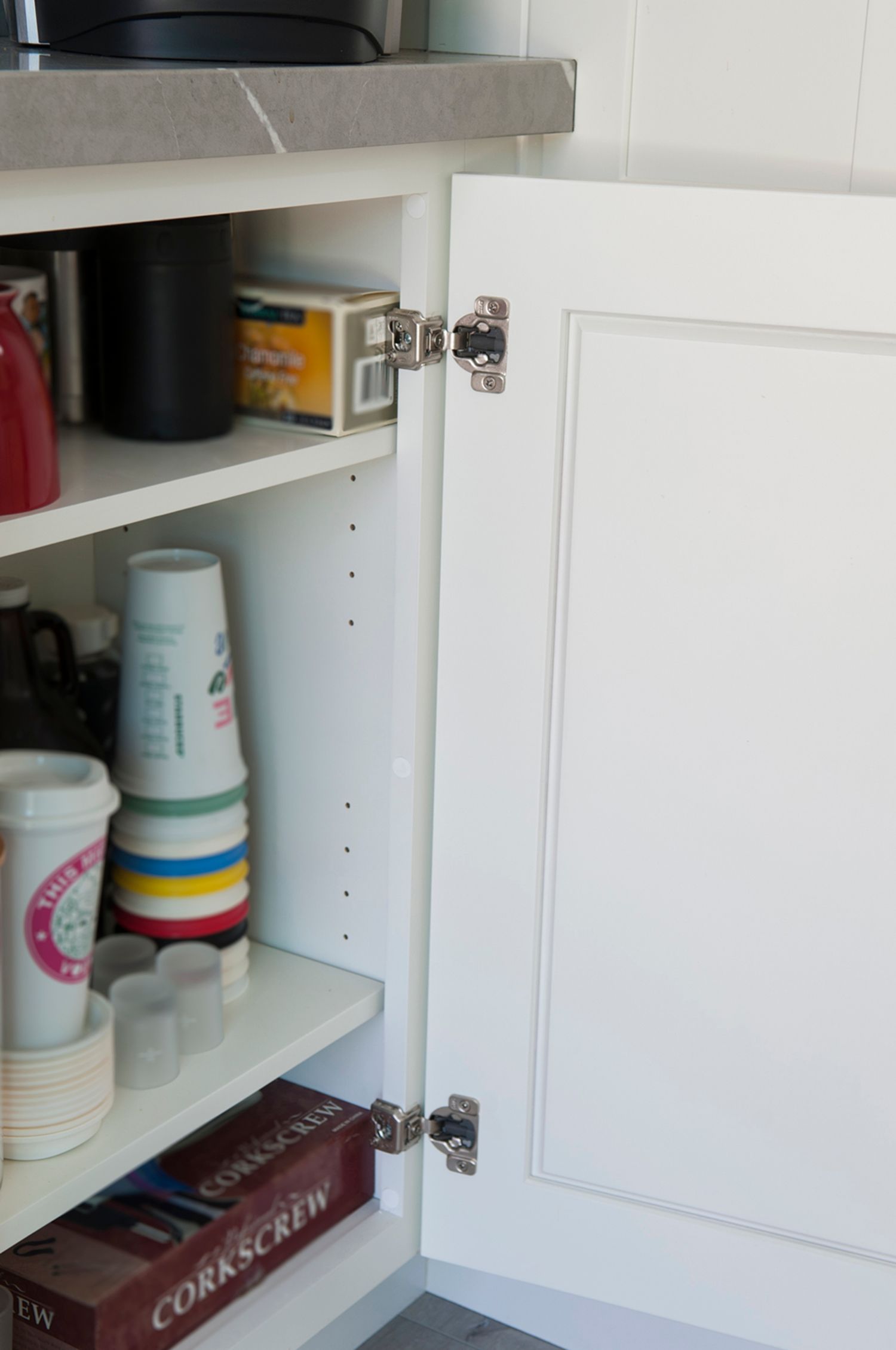 White cabinet with an open door, showing shelves with cups, boxes, and a countertop above.