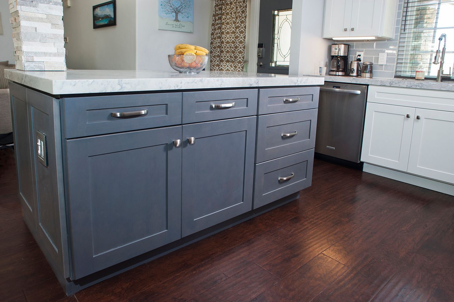 A gray kitchen island with drawers and cabinets, white countertop, in a kitchen with wood floors and a white cabinet.