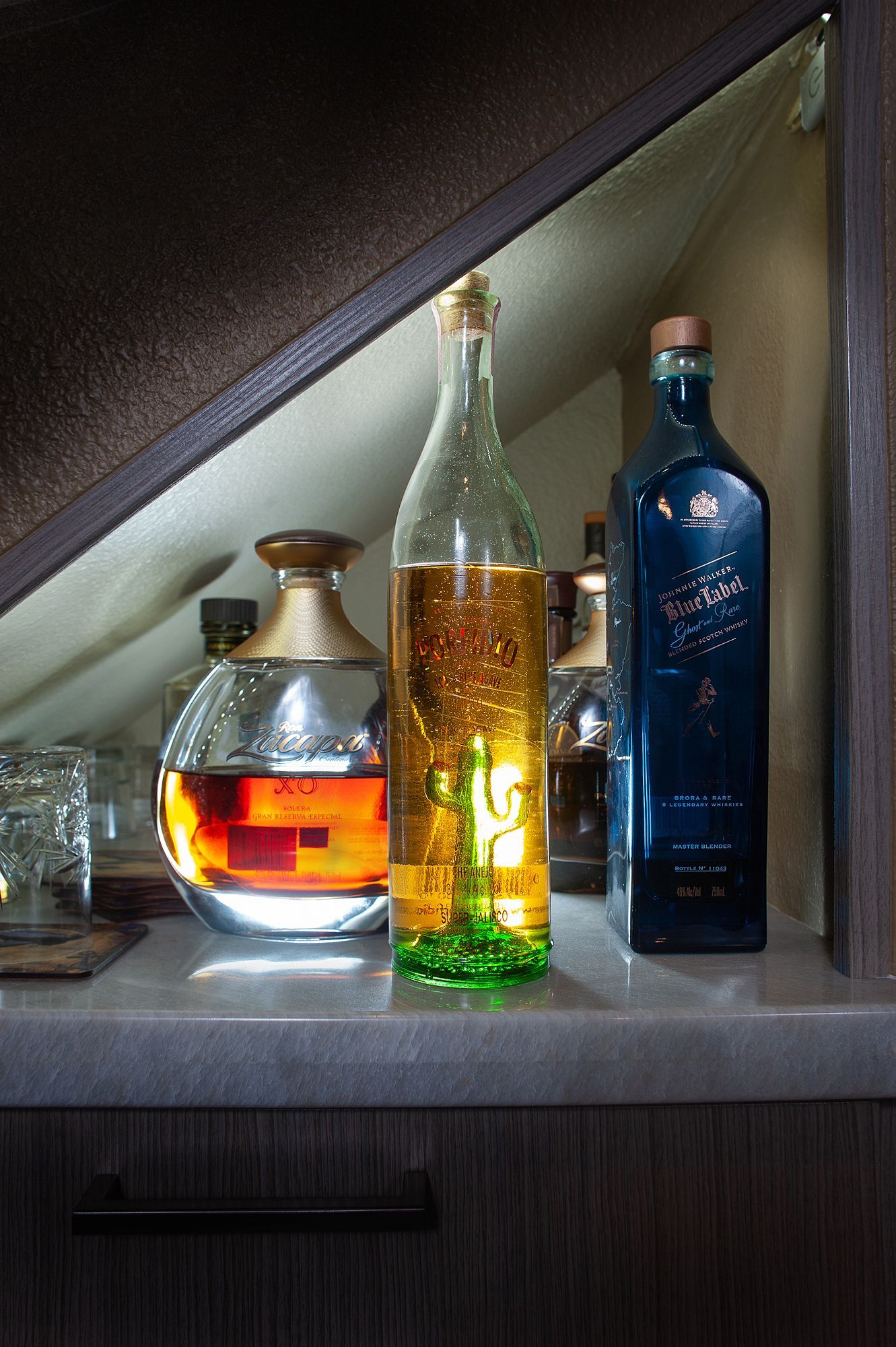 Liquor bottles on a shelf with varying colors, shapes, and sizes, set against a neutral-toned background.