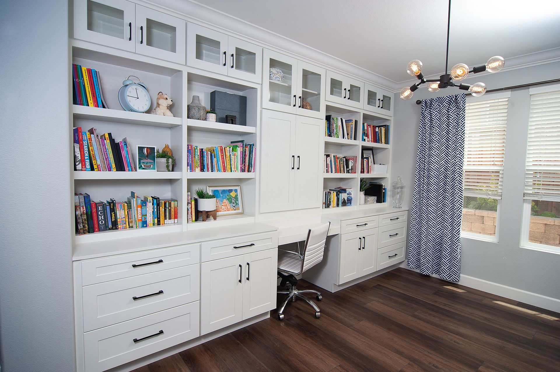 White built-in bookshelves and desk in a home office. Books, decorations, and a desk chair are present. Dark wood floors.