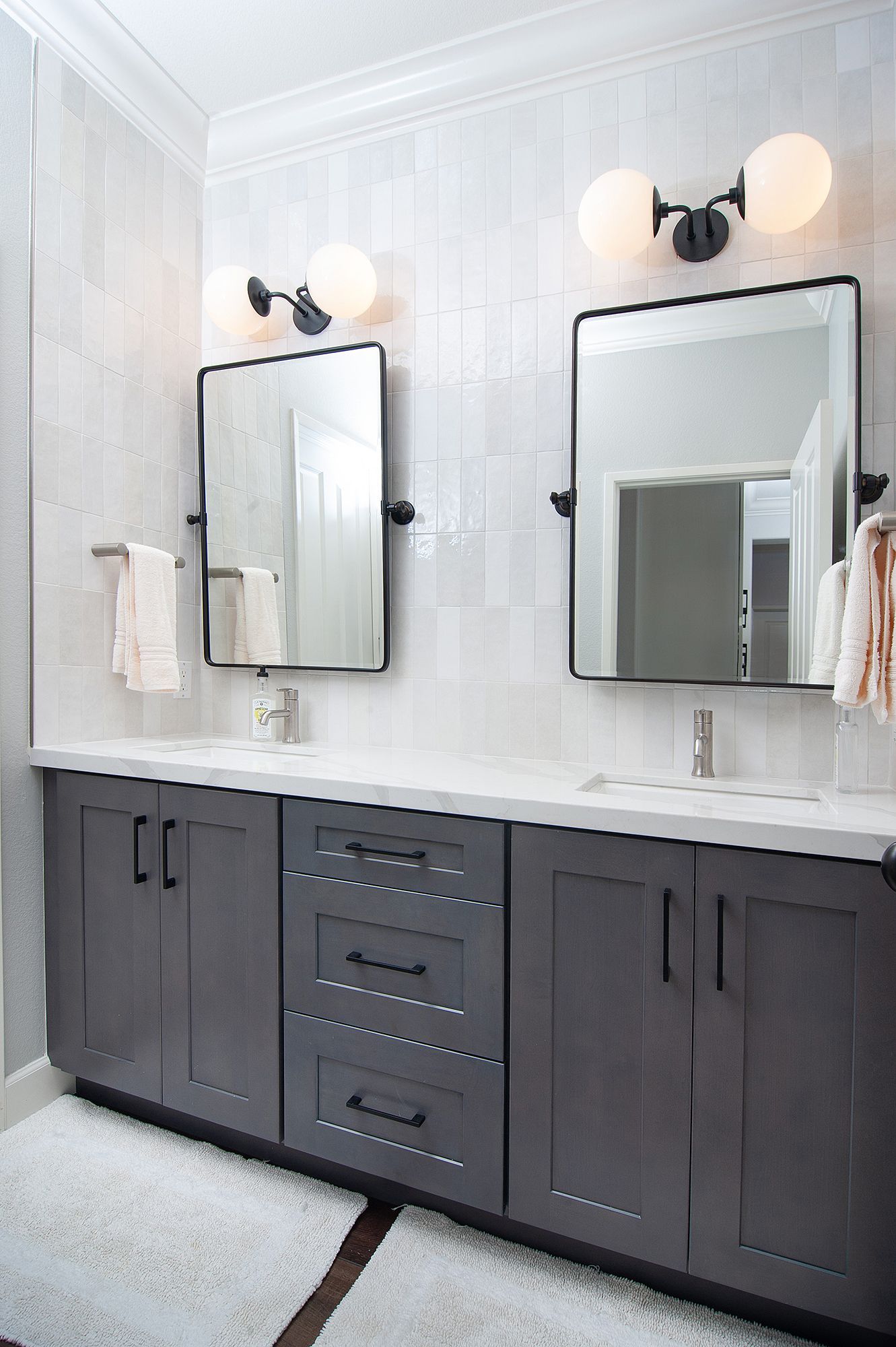 Bathroom with grey vanity, two mirrors, and globe lights against white tiled walls.