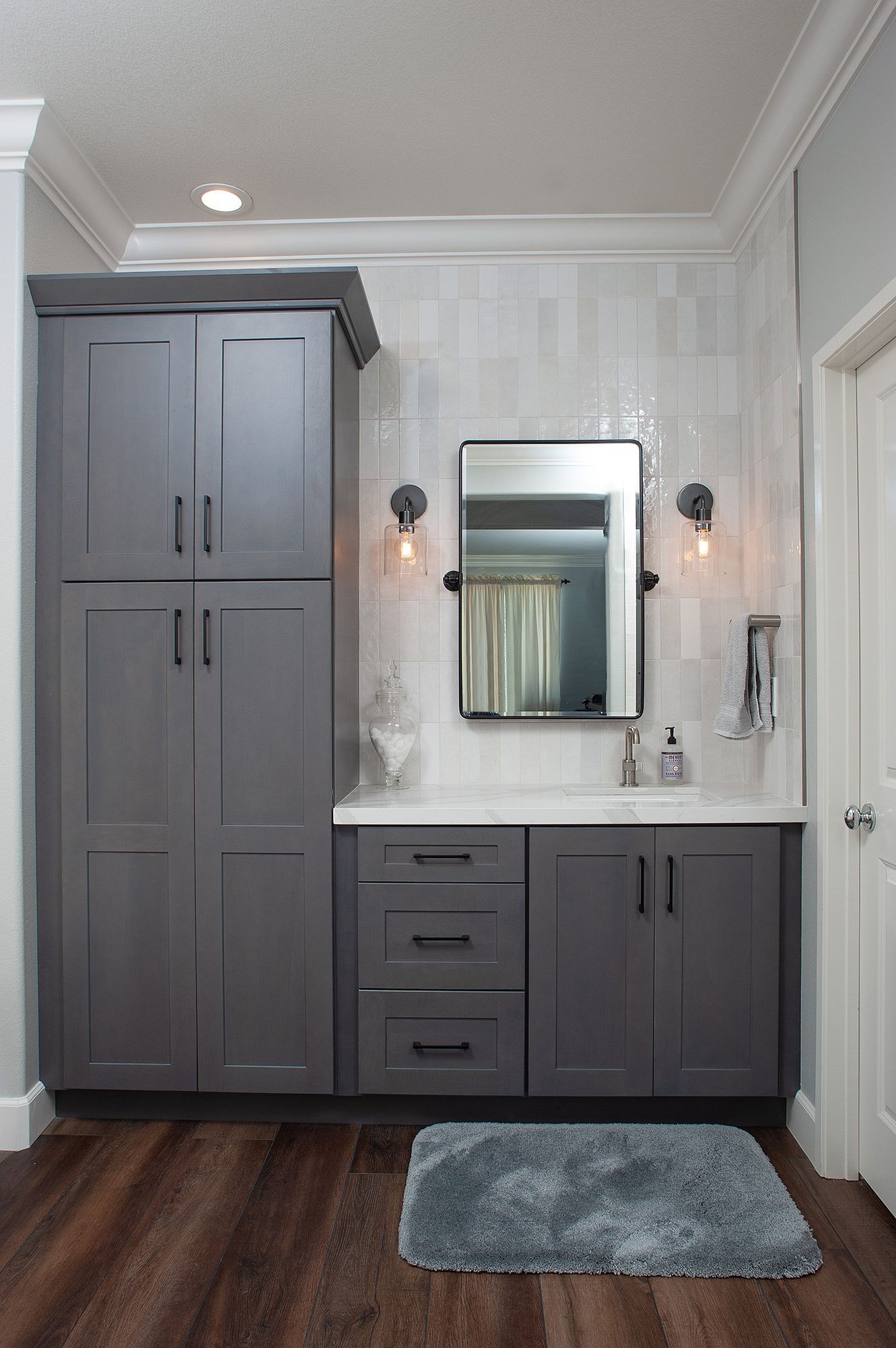 Gray bathroom with tall cabinet, vanity, mirror, and gray rug on wood floor.