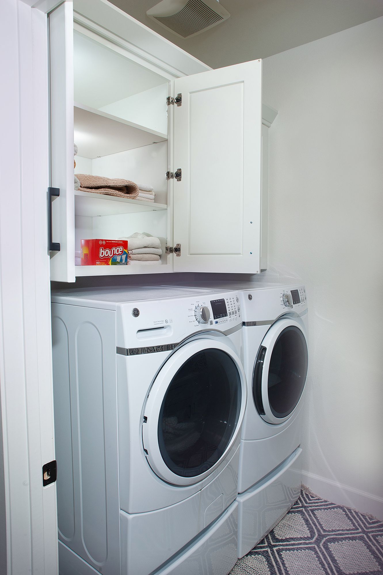White laundry machines stacked under white cabinets in a laundry room; the cabinet door is open.