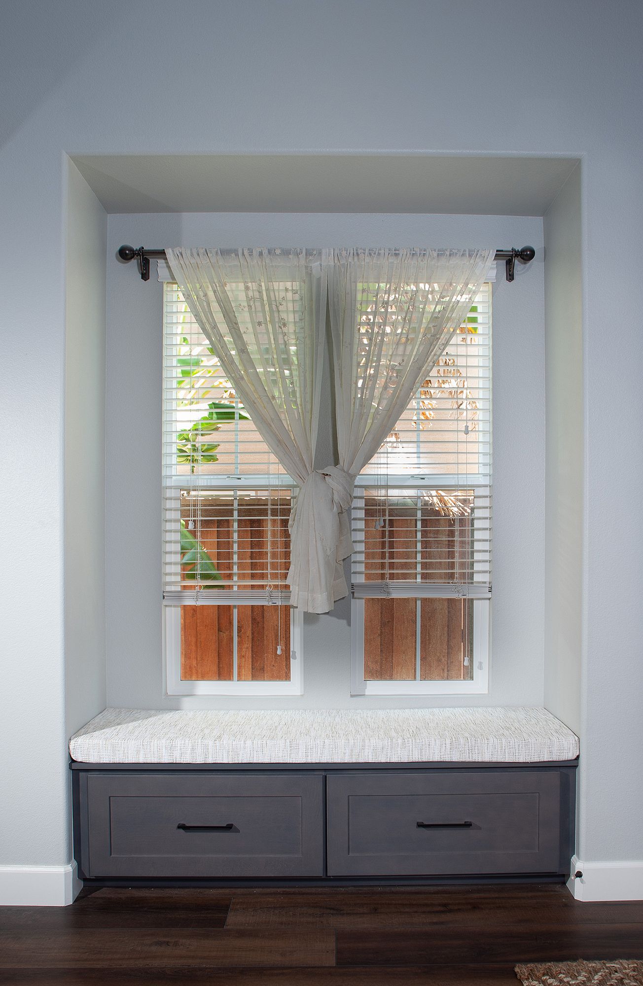 Window seat with gray drawers, white cushion, sheer curtain, and wooden blinds.