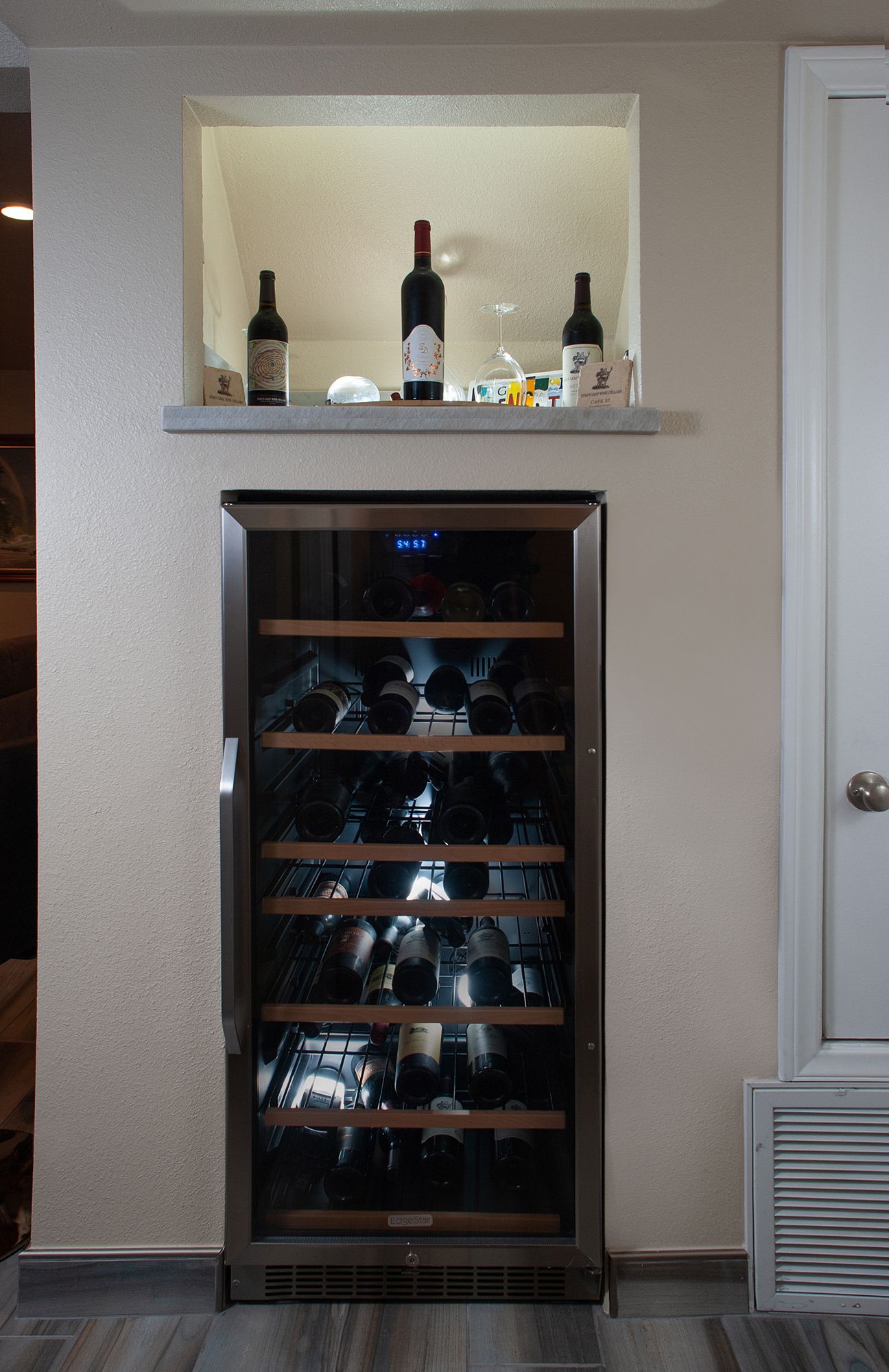 Wine cooler built into a wall niche, with wine bottles displayed above.