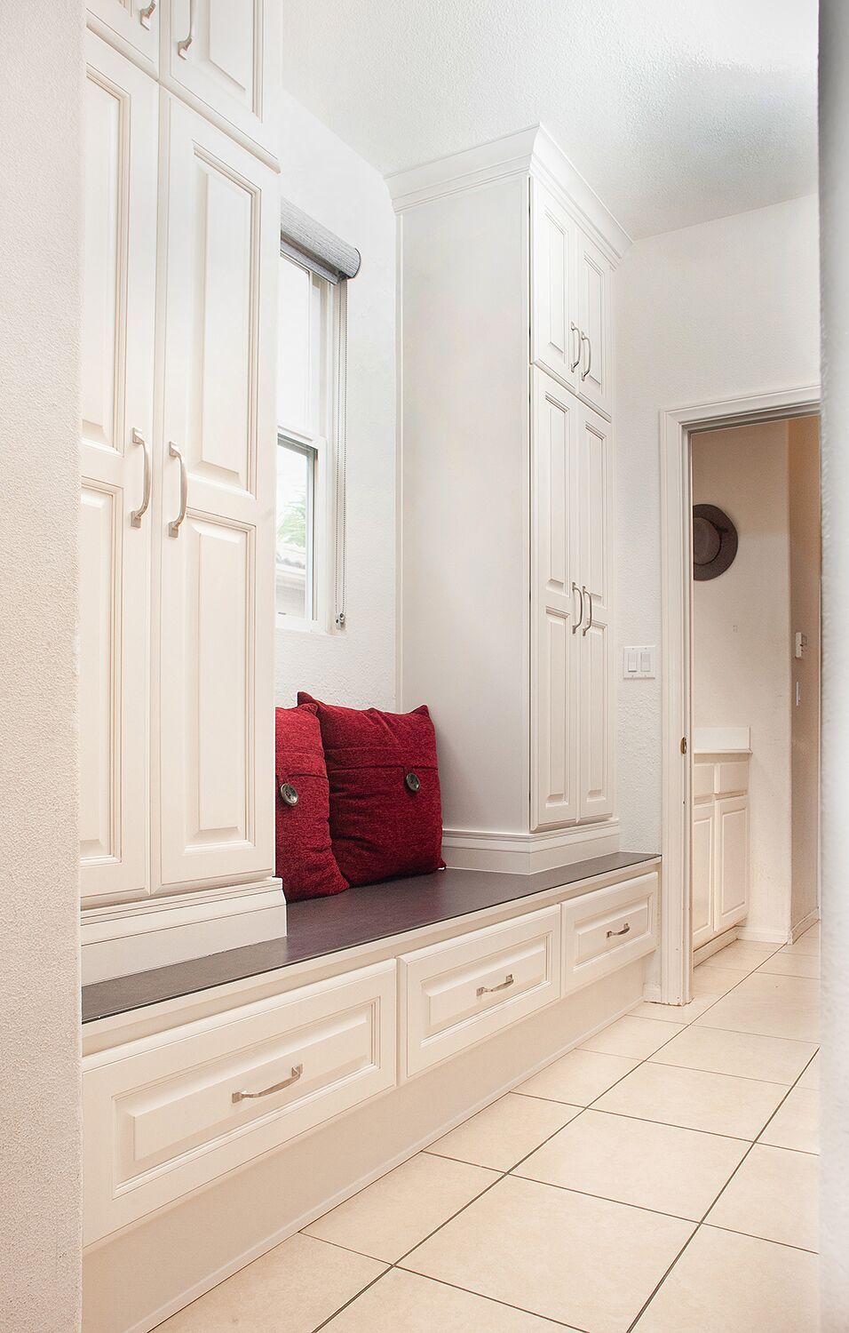 A white built-in bench with drawers and cabinets, red pillows, and a window in a hallway.