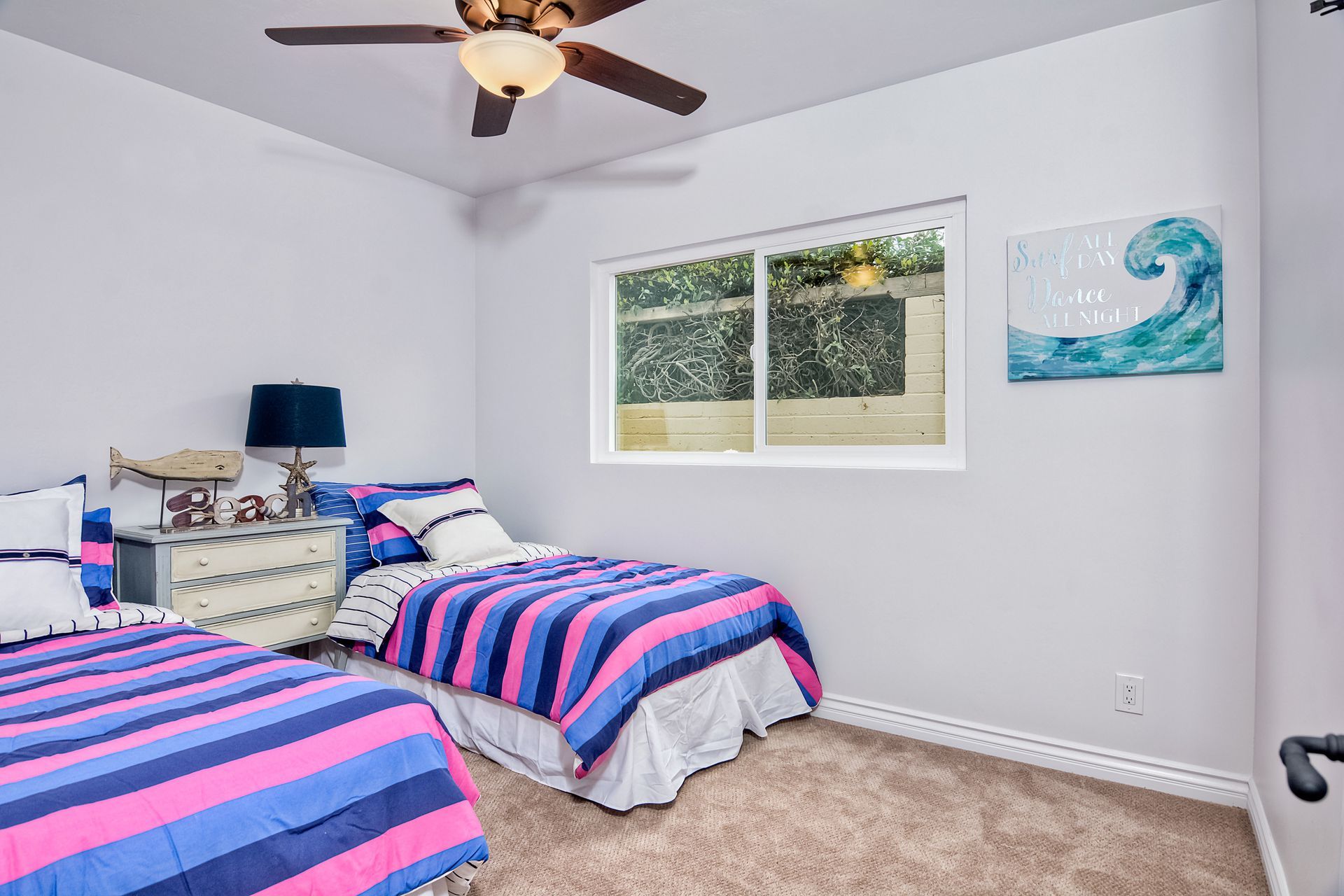 Two twin beds in a bedroom with blue and pink striped comforters. Window with greenery and wave art.