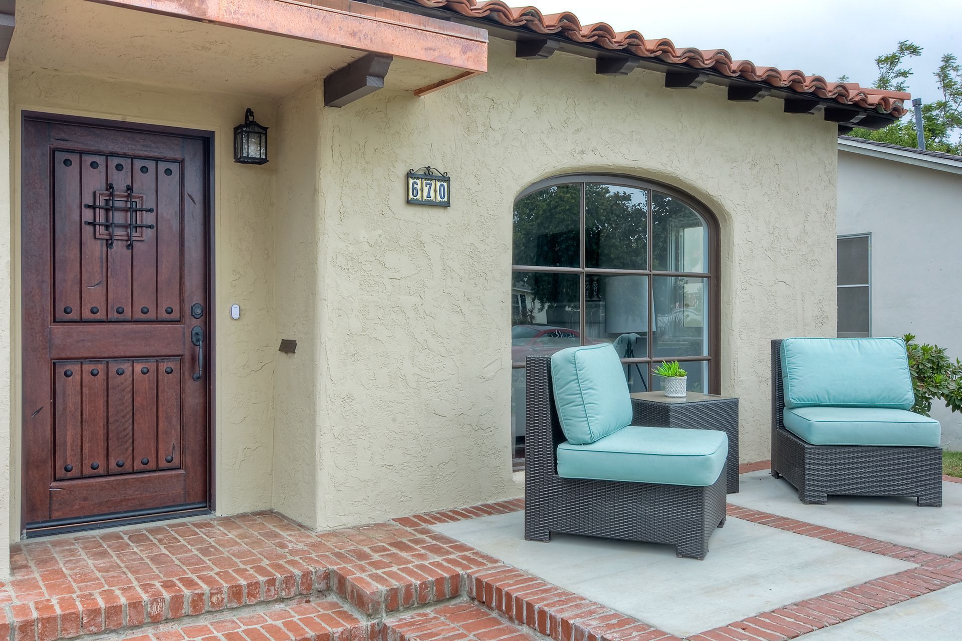 Front exterior of a Spanish-style home with a red brick porch and two blue chairs.