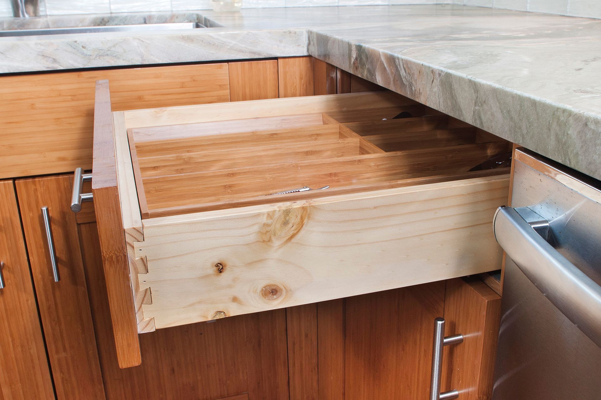 Open wooden kitchen drawer with silverware dividers; drawer is in a wooden cabinet beneath a granite countertop.