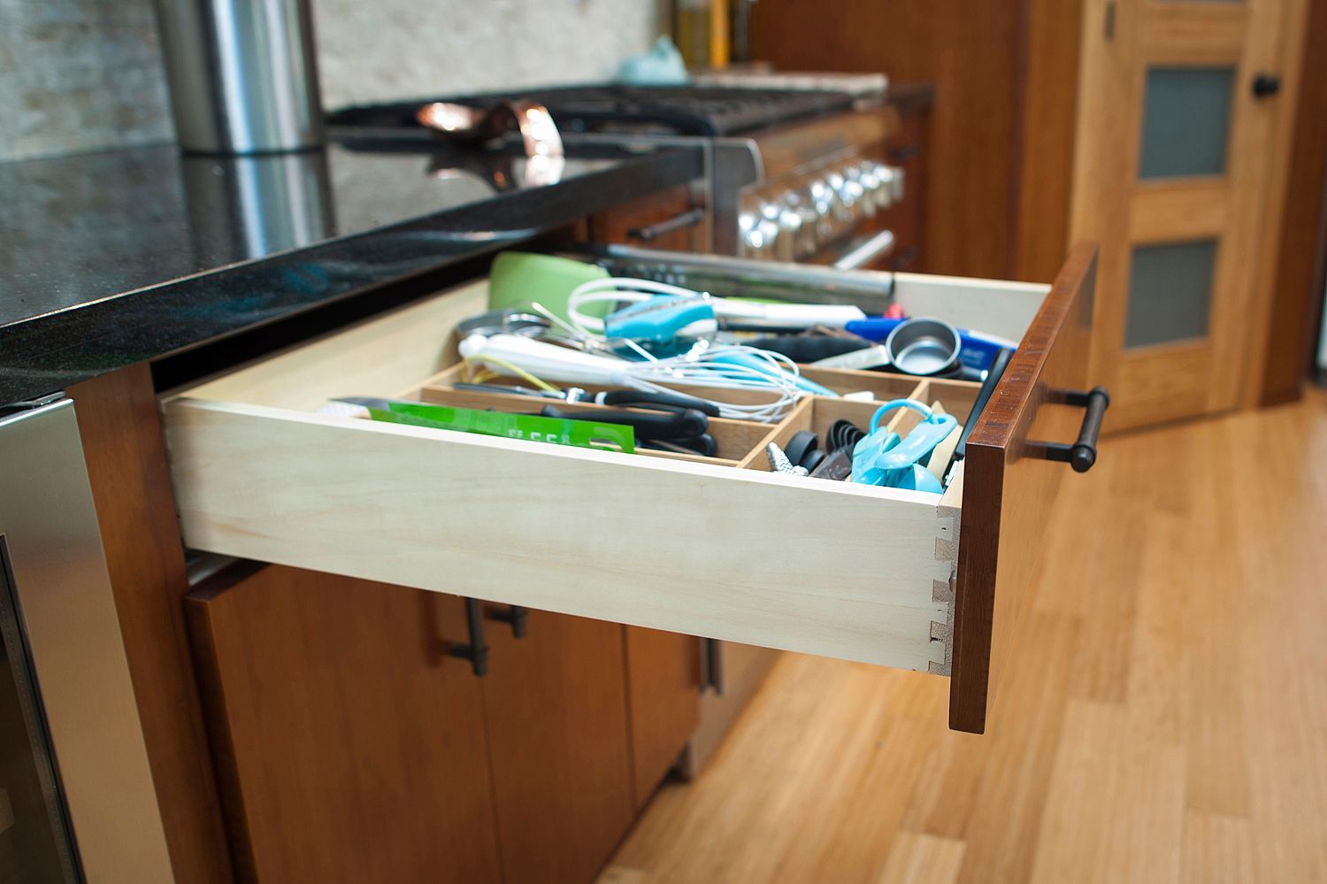 Open kitchen drawer with utensils and dividers. Brown wood cabinets and flooring.