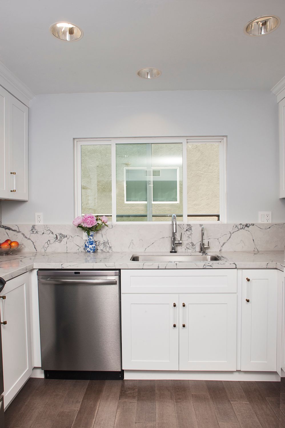 White kitchen with stainless steel dishwasher, sink, and marble countertops.