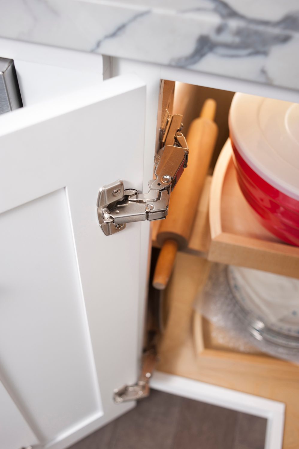White kitchen cabinet door open, revealing a pull-out tray with a rolling pin and a red bowl.