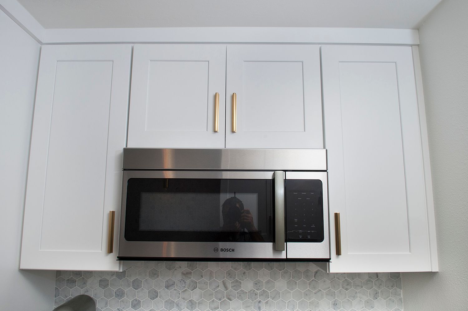 White kitchen cabinets above a stainless steel microwave; gold handles; white and gray backsplash.