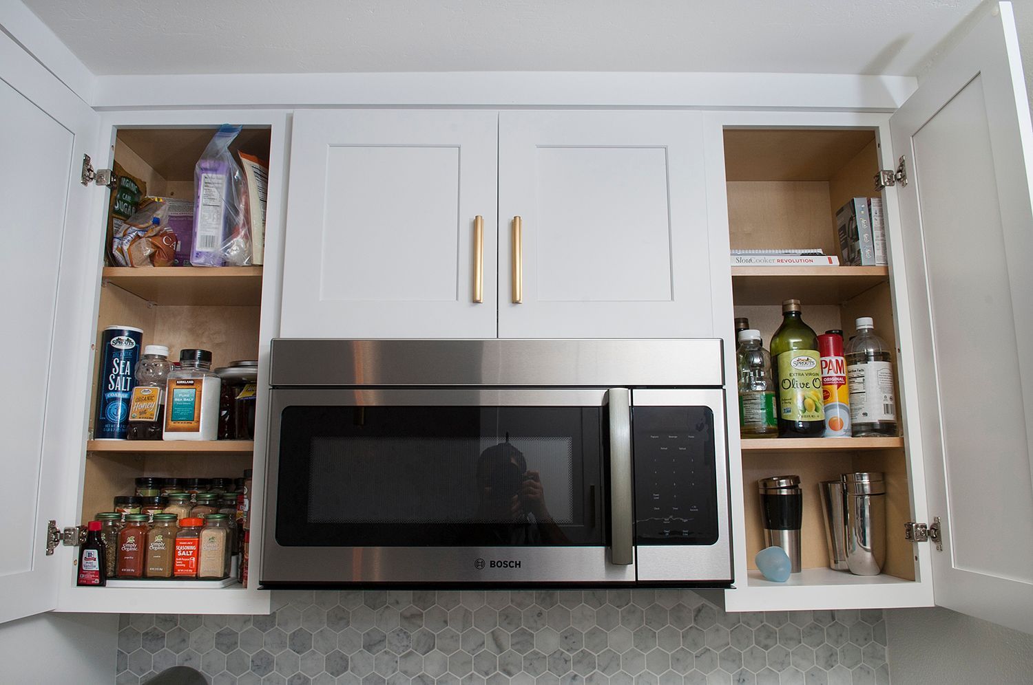 Kitchen cabinet with open doors, above a microwave. Inside are spices, jars, and bottles.