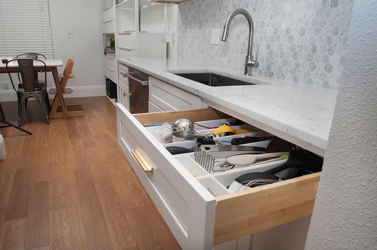 Kitchen drawer open, revealing utensils and storage. White cabinets, wood floor, and sink visible.