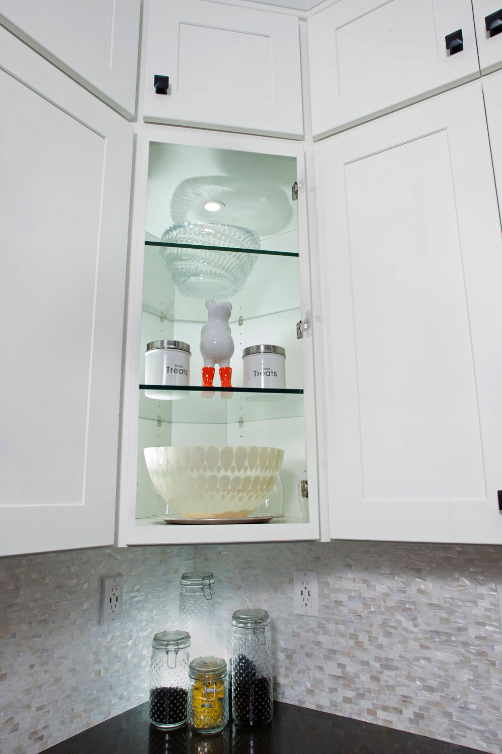 Corner kitchen cabinet with glass shelves, displaying dishware and jars.