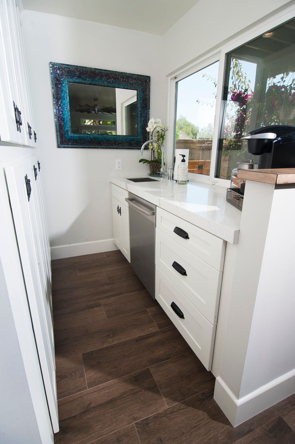 White kitchenette with dishwasher, sink, and mirror next to a sliding glass door. Dark wood floor.